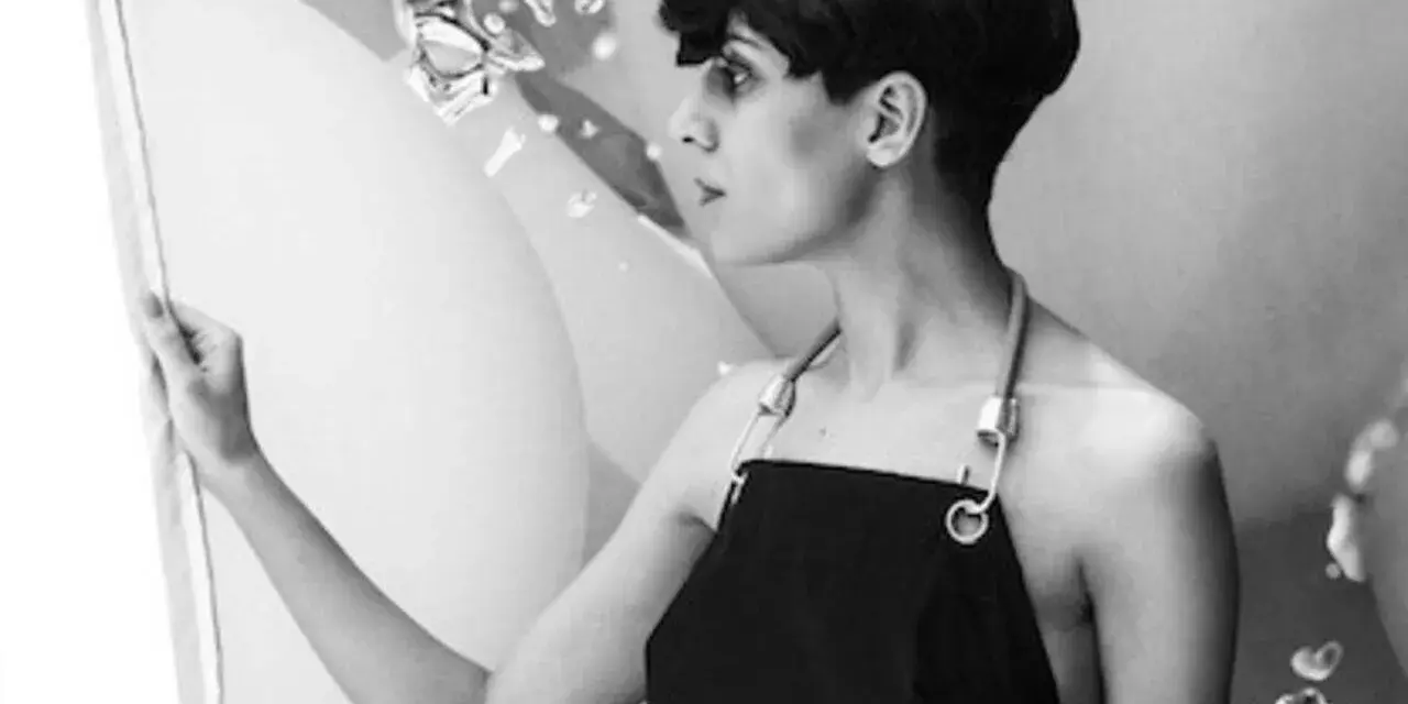 Fatima Al Qadiri, artiste à la coupe courte, porte un tablier noir, regardant une œuvre abstraite.