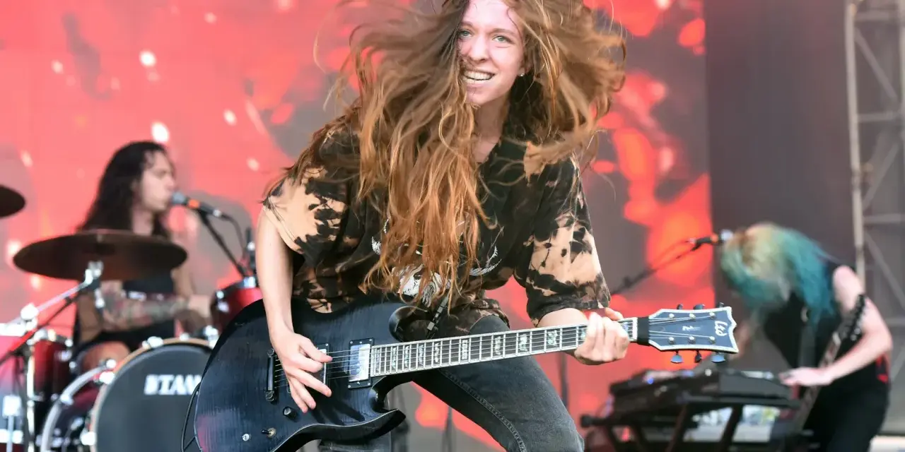 Reba Meyers, guitariste au look sauvage, fait vibrer sa guitare électrique sur scène, cheveux au vent, devant un fond rougeoyant.
