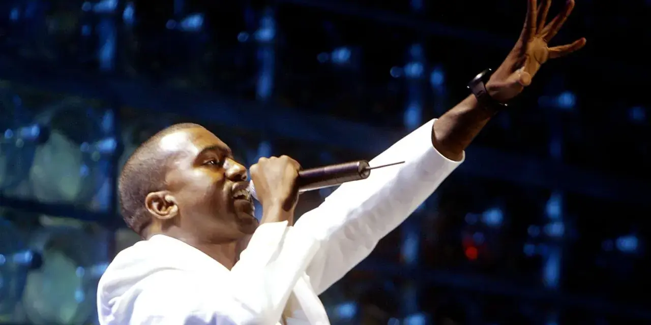 Kanye West, aux origines de sa carrière, chante sur scène, micro à la main, bras levé.