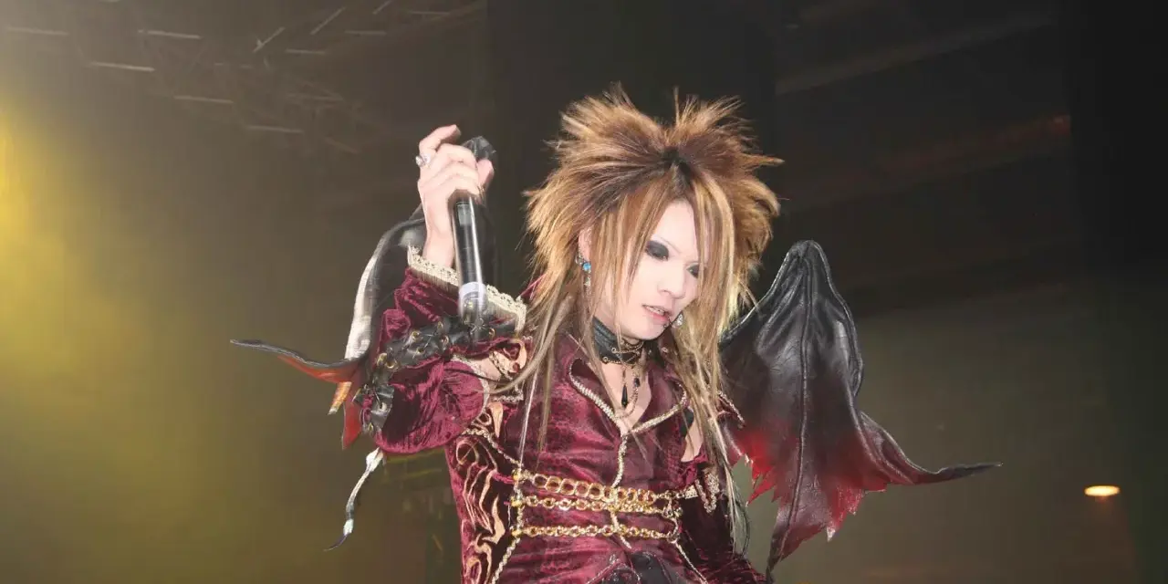 Un membre de X Japan, cheveux ébouriffés, maquillage prononcé, costume sombre et ailes de démon, chante sur scène.