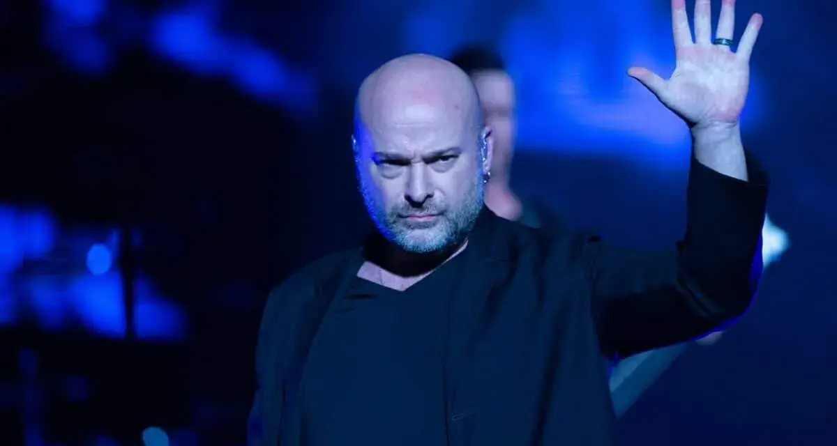 Le chanteur Disturbed, chauve et barbu, lève la main sur scène sous des lumières bleues intenses.