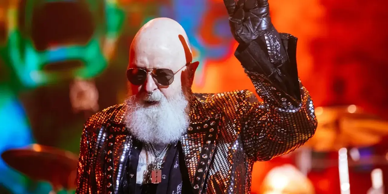 Rob Halford, un des membres de Judas Priest, fait le signe des cornes sur scène, vêtu d'une veste scintillante.
