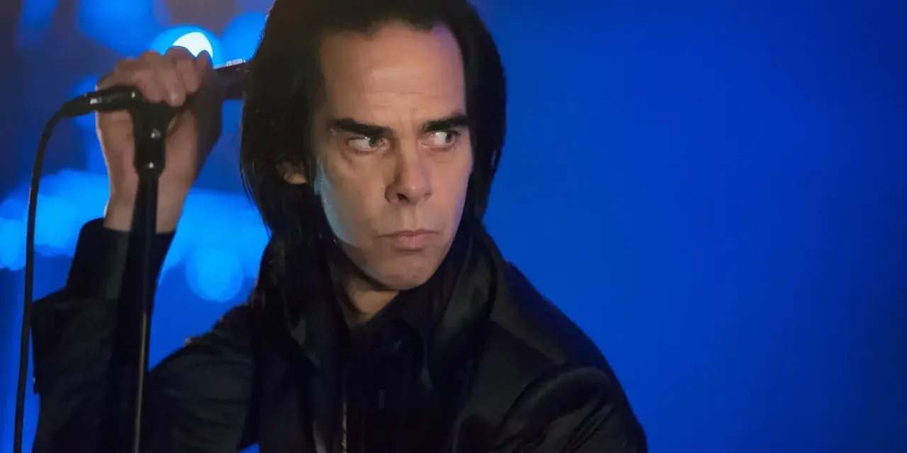 Nick Cave, chanteur du groupe australien des années 80, sur scène, micro à la main, regard intense sur fond bleu.