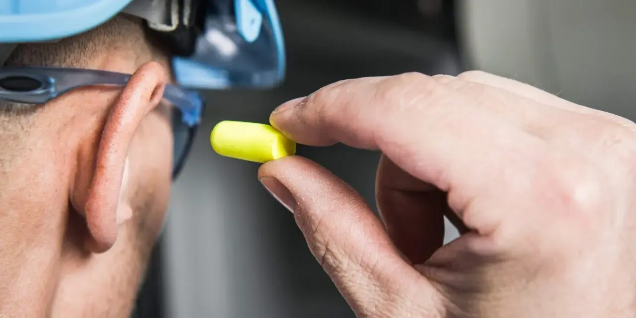 Main tenant, casque bleu, lunettes de protection et bouchon d'oreille jaune pour se protéger d'un bruit de 120 décibels équivalent.