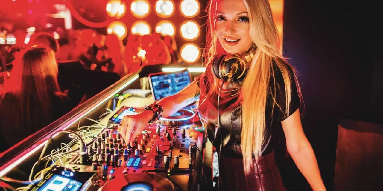 Une dj femme connue, aux cheveux blonds, mixe dans une boîte de nuit. La foule est en arrière-plan, éclairée par des lumières rouges.