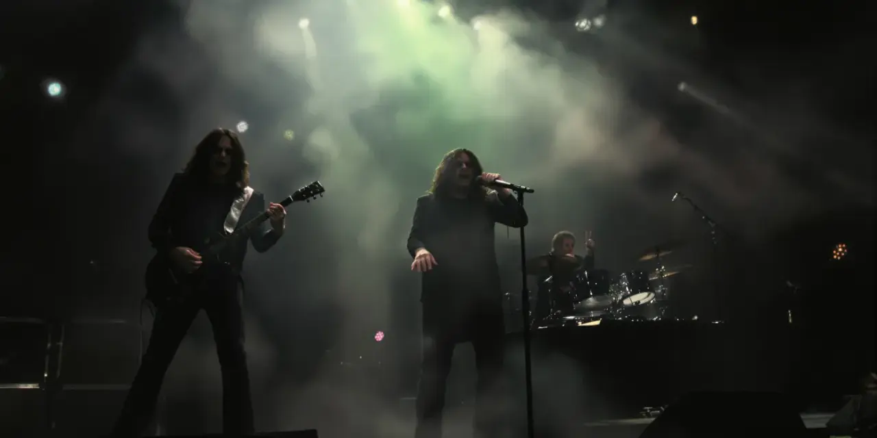 Un groupe de metal sur scène, avec un guitariste à gauche, un chanteur au centre, et un batteur à droite. La scène est enfumée, éclairée par des projecteurs.