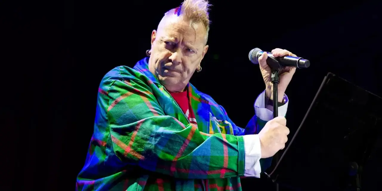 Le chanteur des Sex Pistols, John Lydon, avec sa crête iconique, porte une veste à carreaux écossais colorée et tient un microphone.