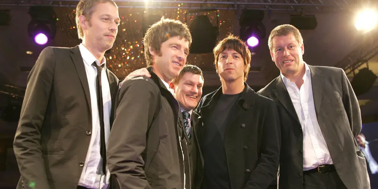 Cinq hommes, dont le groupe Oasis, posent ensemble sur scène, souriants sous les lumières.