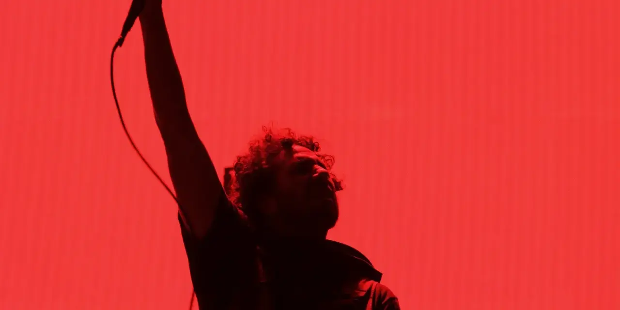 Chanteur de Rage Against the Machine, micro levé, sur fond rouge vif. L'énergie de leurs albums en live.