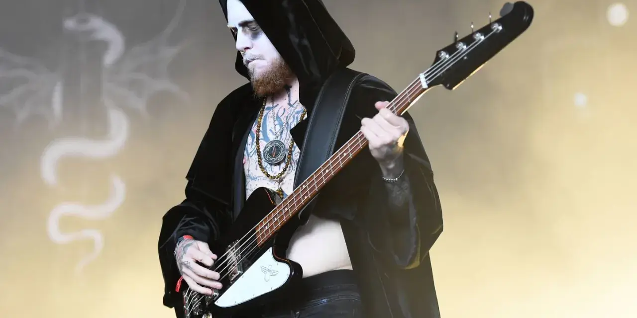 Le bassiste d'un groupe metal connu, maquillé et tatoué, joue sur scène, drapé dans une cape noire.