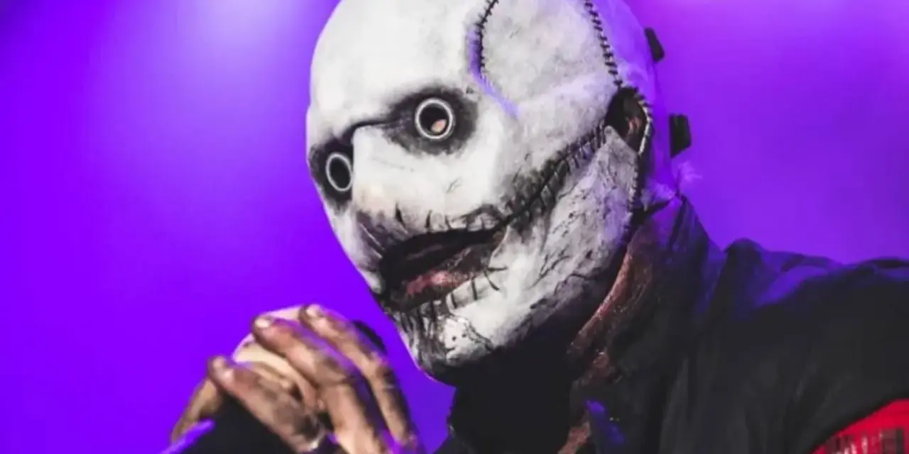 Le chanteur Slipknot, masqué et terrifiant, tient un micro sur scène sous une lumière violette.