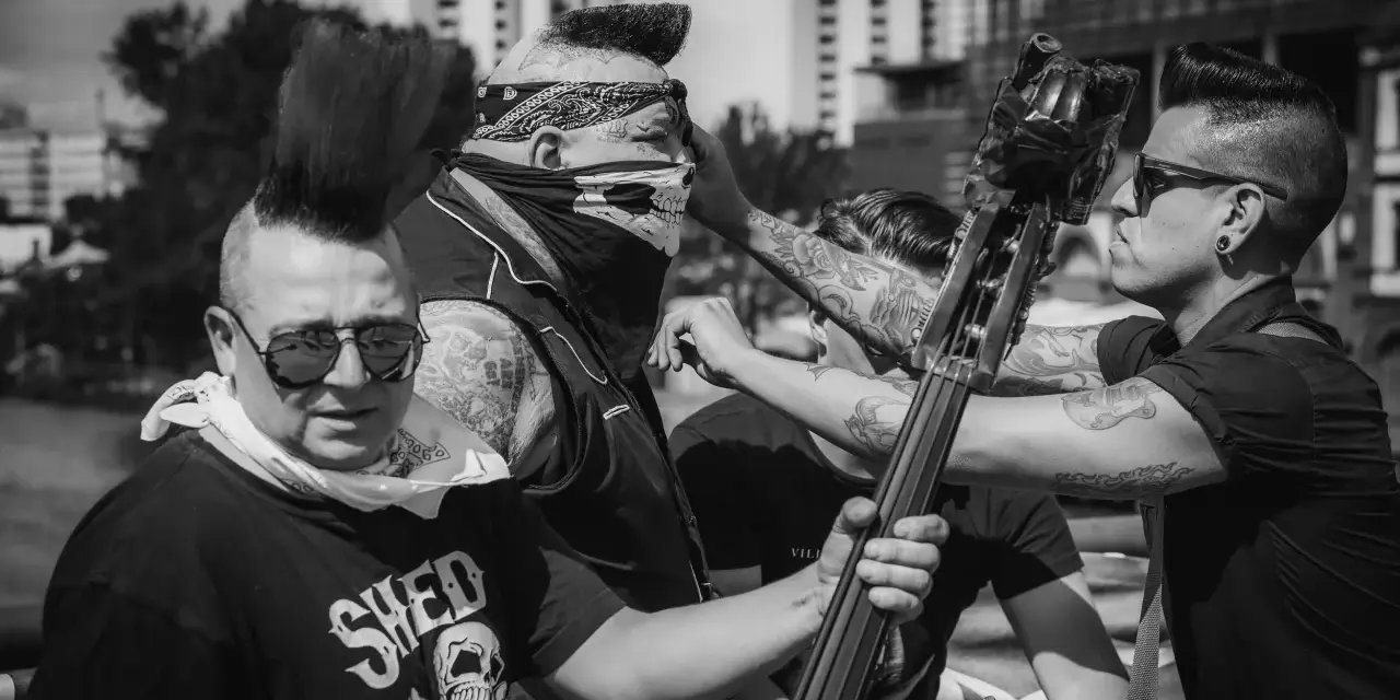 Quatre musiciens au look punk, dont un avec un bandana et un masque de crâne, se préparent à jouer. Un d'eux tient une contrebasse, prêt à faire un mad sin.