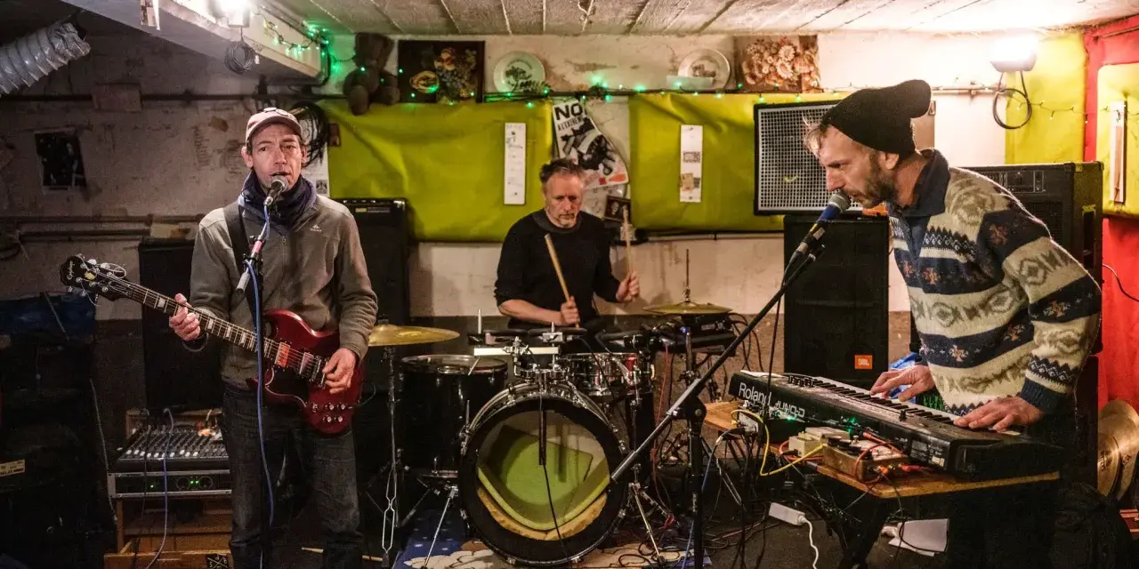 Trois musiciens jouent dans un local sombre, éclairé par des guirlandes. Un guitariste, un batteur et un claviériste, oi boys !