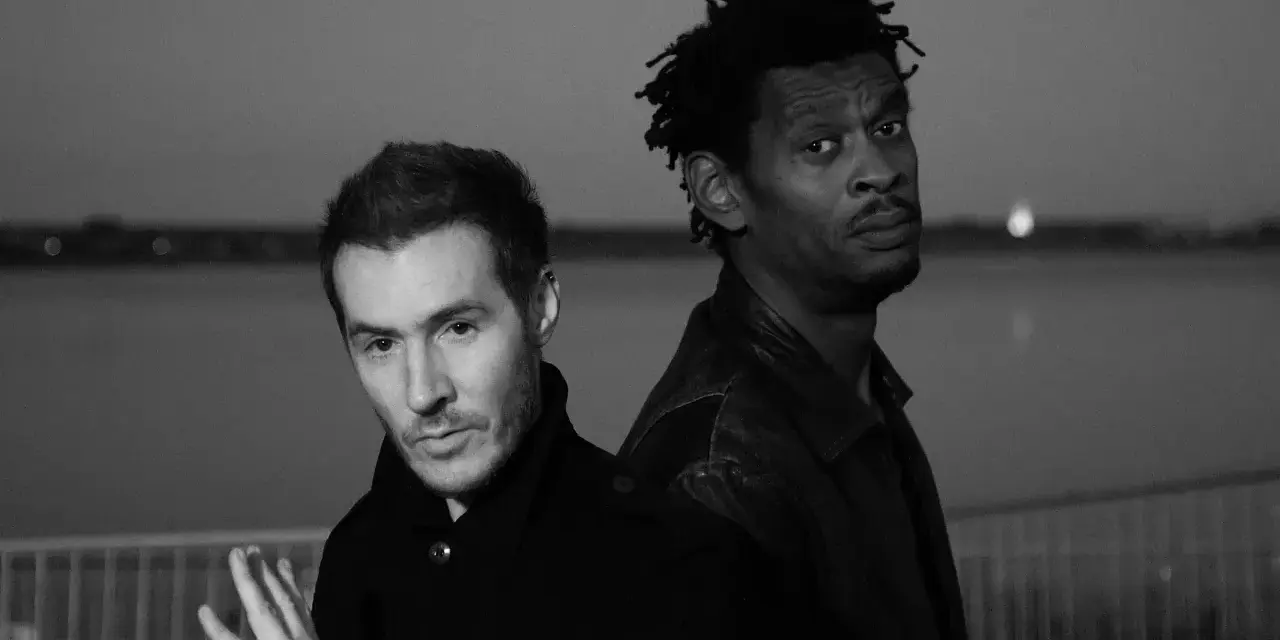 Les membres de Massive Attack, 3D et Daddy G, posent dos à dos, regardant dans des directions opposées, dans une ambiance sombre et contemplative.