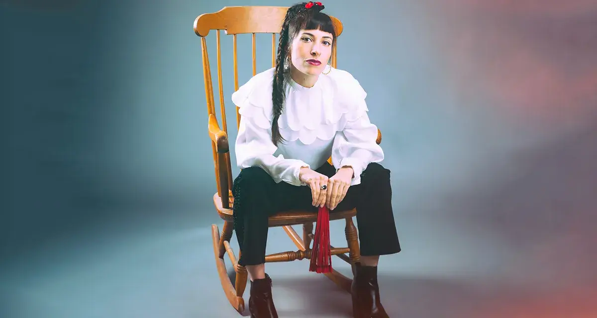 Scout Niblett, assise sur un fauteuil à bascule, porte une blouse blanche à volants et un pantalon noir. Elle tient un pompon rouge.