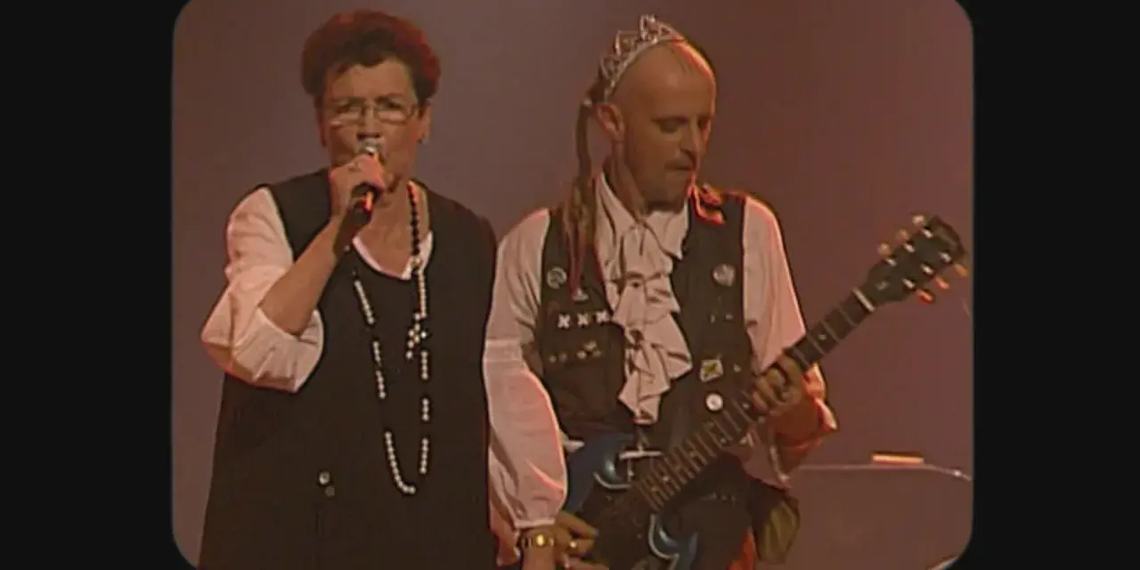 Une chanteuse et un guitariste, peut-être les ramoneurs de menhirs, sur scène. L'homme porte une couronne et une tenue excentrique.