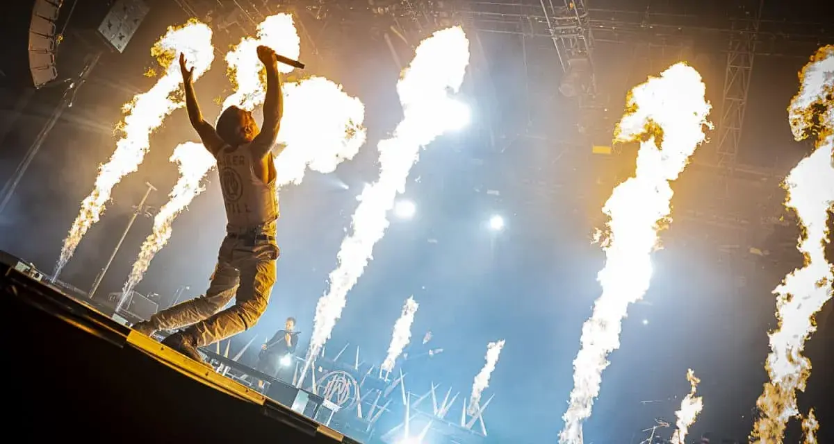 Un chanteur de groupe hard rock des années 2000, micro en main, saute sur scène entouré de jets de flammes spectaculaires.