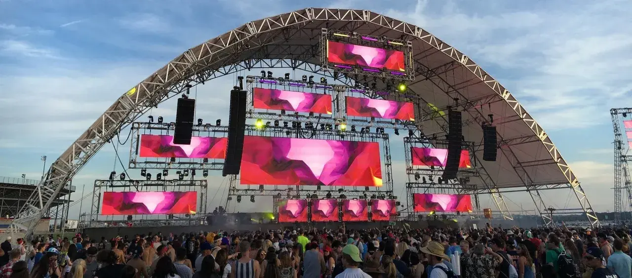Scène de festival avec écrans LED diffusant des visuels abstraits roses et violets. Une foule danse sous la structure, attendant leur artiste deep house préféré.