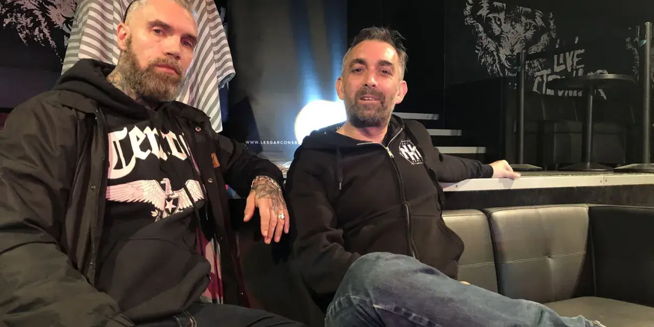 Deux hommes, dont un possible chanteur de Mass Hysteria, posent sur un canapé. L'un porte un sweat à capuche noir avec un aigle.