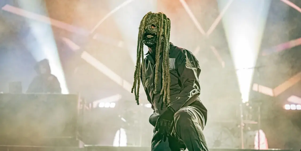 Un membre de Slipknot, masqué et en combinaison, sur scène sous des projecteurs.