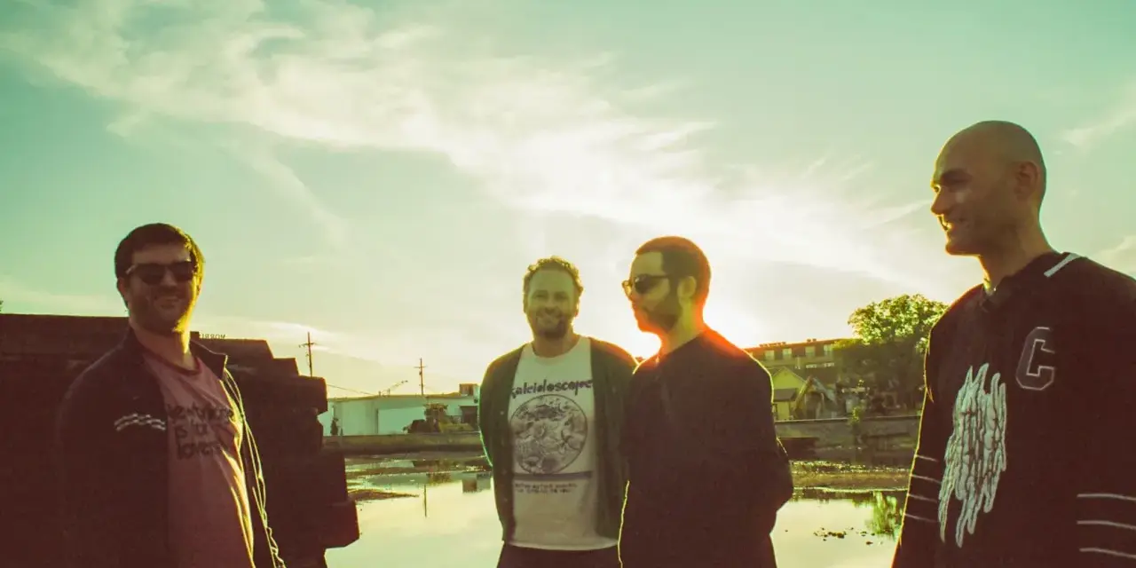 Quatre hommes posent devant un ciel clair, comme un chat pile. L'un porte un t-shirt "kaleidoscope".