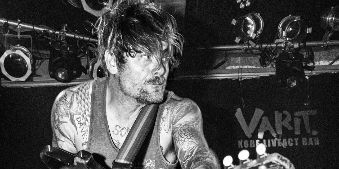 Un guitariste tatoué, cheveux en bataille, sous les projecteurs du VART. Kobe Liveact Bar.