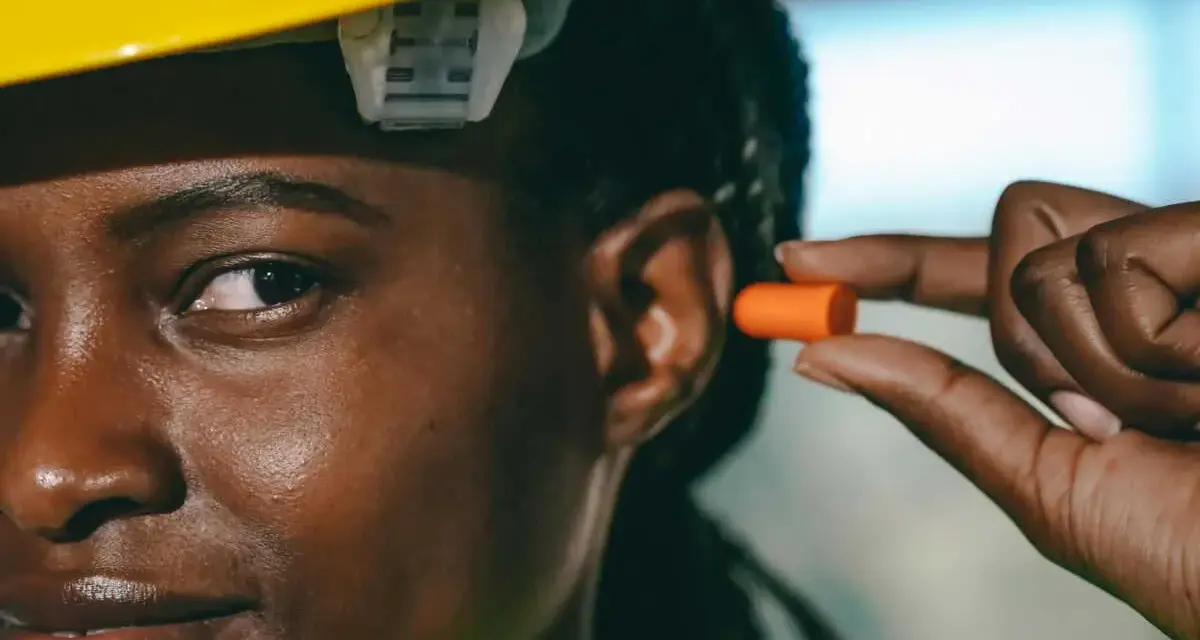Une femme porte un casque jaune et s'apprête à insérer un bouchon d'oreille orange pour éviter le sifflement.