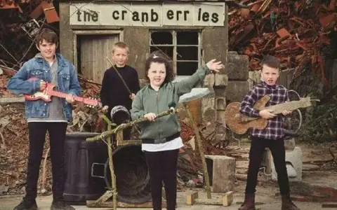 Quatre enfants jouent de la musique devant un panneau "the cranberries". L'un tient une guitare rose, un autre une guitare en bois.