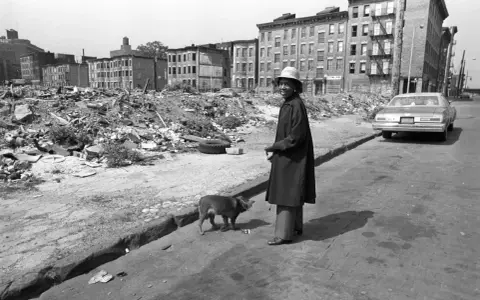 Une femme et son chien devant des immeubles délabrés et des décombres.
