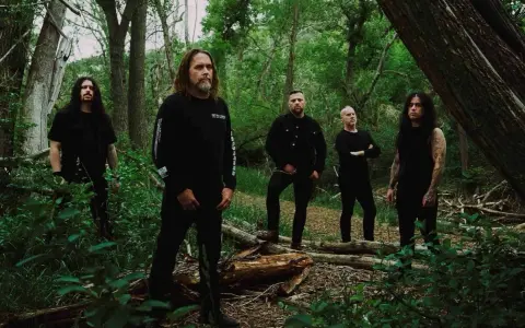 Cinq hommes en noir posent dans une forêt. Le groupe, qui pourrait être Cattle Decapitation, se tient parmi les arbres et la végétation luxuriante.