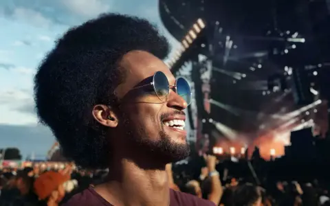 Un homme souriant avec des lunettes de soleil profite d'un concert en plein air. Il porte des bouchons d'oreilles, se demandant pourquoi pas de bouchon concert pour protéger son audition.