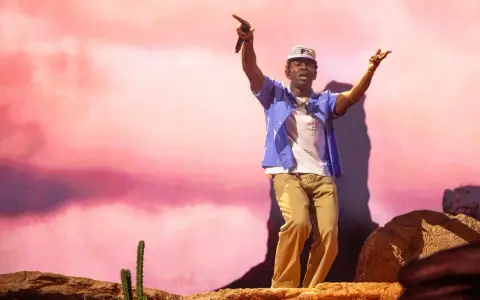 Tyler Okonma, micro en main, danse sur scène devant un décor rose pastel. Il porte une casquette, une chemise bleue ouverte sur un t-shirt blanc et un pantalon beige.