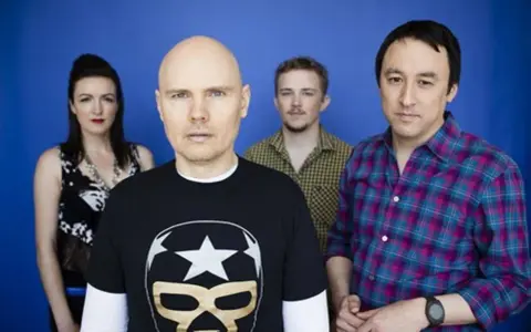 Les membres de The Smashing Pumpkins posent devant un fond bleu. Billy Corgan porte un t-shirt noir avec un masque de lucha.