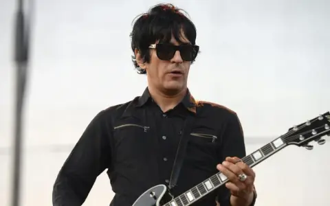 Un des membres de Eagles of Death Metal, guitariste en chemise noire et lunettes de soleil, joue sur scène.