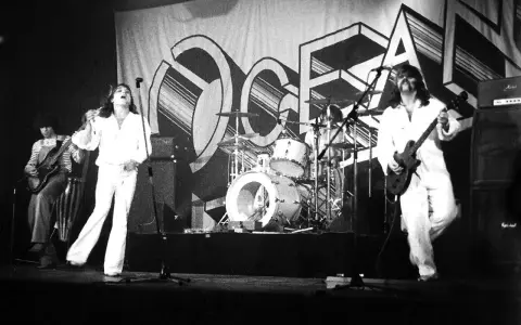 Le groupe Trust en concert, avec le chanteur en blanc au centre, un guitariste à gauche et un autre guitariste à droite. La scène est décorée d'une grande bannière avec le logo du groupe.