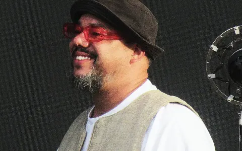 Un chanteur soul souriant, portant un chapeau et des lunettes rouges, se tient devant un microphone.