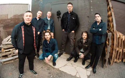 Les membres de Dropkick Murphys posent devant un mur sombre, certains accroupis, d'autres debout, prêts pour leur prochain concert.