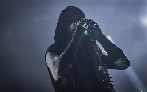 Chanteur de Dimmu Borgir, visage encapuchonné et maquillé, chantant dans un micro, ambiance sombre et enfumée.