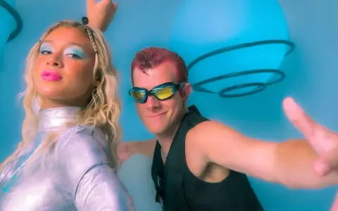 Un groupe hollandais des années 90, avec une femme au maquillage bleu vif et un homme aux lunettes futuristes, pose devant des sphères lumineuses bleues.