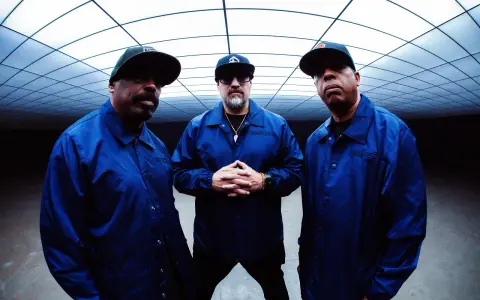 Les membres de Cypress Hill, B-Real, Sen Dog et DJ Muggs, posent ensemble, portant des vestes bleues assorties sous un plafond géométrique.