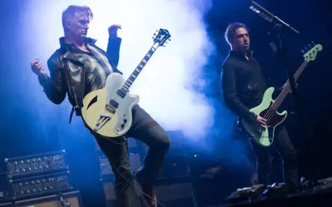 Deux membres de Queens of the Stone Age en pleine performance, l'un jouant de la guitare, l'autre de la basse, sous des éclairages bleus intenses.