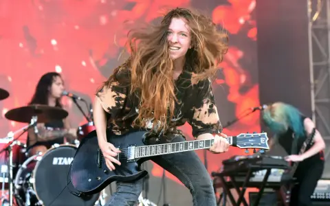Reba Meyers, guitariste au look sauvage, fait vibrer sa guitare électrique sur scène, cheveux au vent, devant un fond rougeoyant.