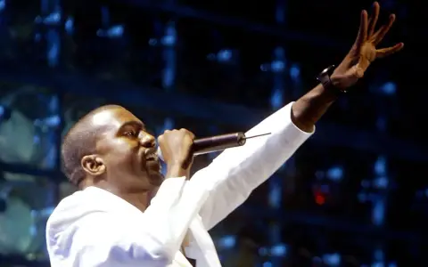 Kanye West, aux origines de sa carrière, chante sur scène, micro à la main, bras levé.