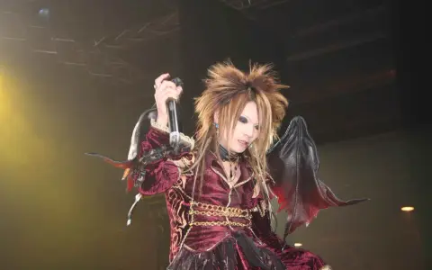 Un membre de X Japan, cheveux ébouriffés, maquillage prononcé, costume sombre et ailes de démon, chante sur scène.