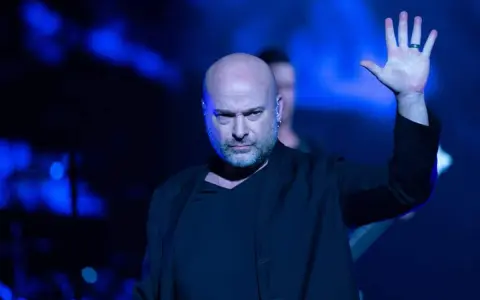 Le chanteur Disturbed, chauve et barbu, lève la main sur scène sous des lumières bleues intenses.