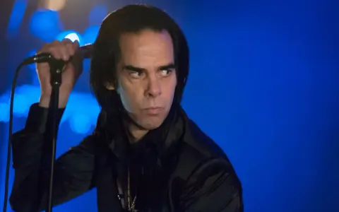 Nick Cave, chanteur du groupe australien des années 80, sur scène, micro à la main, regard intense sur fond bleu.