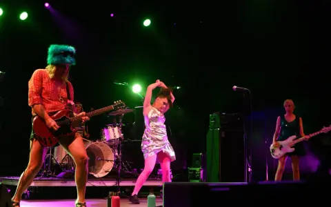 Sur scène, le groupe Bikini Kill enflamme le public. Une guitariste porte un chapeau vert fluo, une autre joue de la basse, et une troisième danse avec énergie.