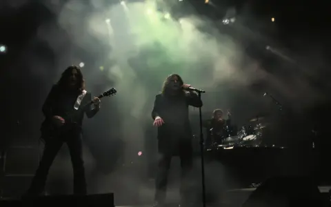 Un groupe de metal sur scène, avec un guitariste à gauche, un chanteur au centre, et un batteur à droite. La scène est enfumée, éclairée par des projecteurs.