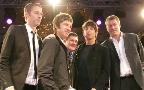 Cinq hommes, dont le groupe Oasis, posent ensemble sur scène, souriants sous les lumières.