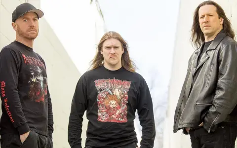 Trois hommes posent devant un mur. L'un porte un t-shirt avec une image de fœtus mourant.